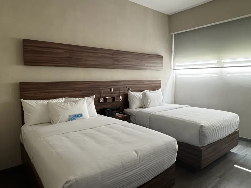 Habitación con dos camas de ropa blanca, cabeceras con veta de madera, lámparas de lectura y piso oscuro